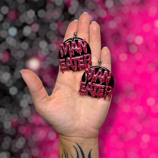 Hand holding a pair of Man Eater drippy glittery resin round dangle earrings on pink and black blurred background