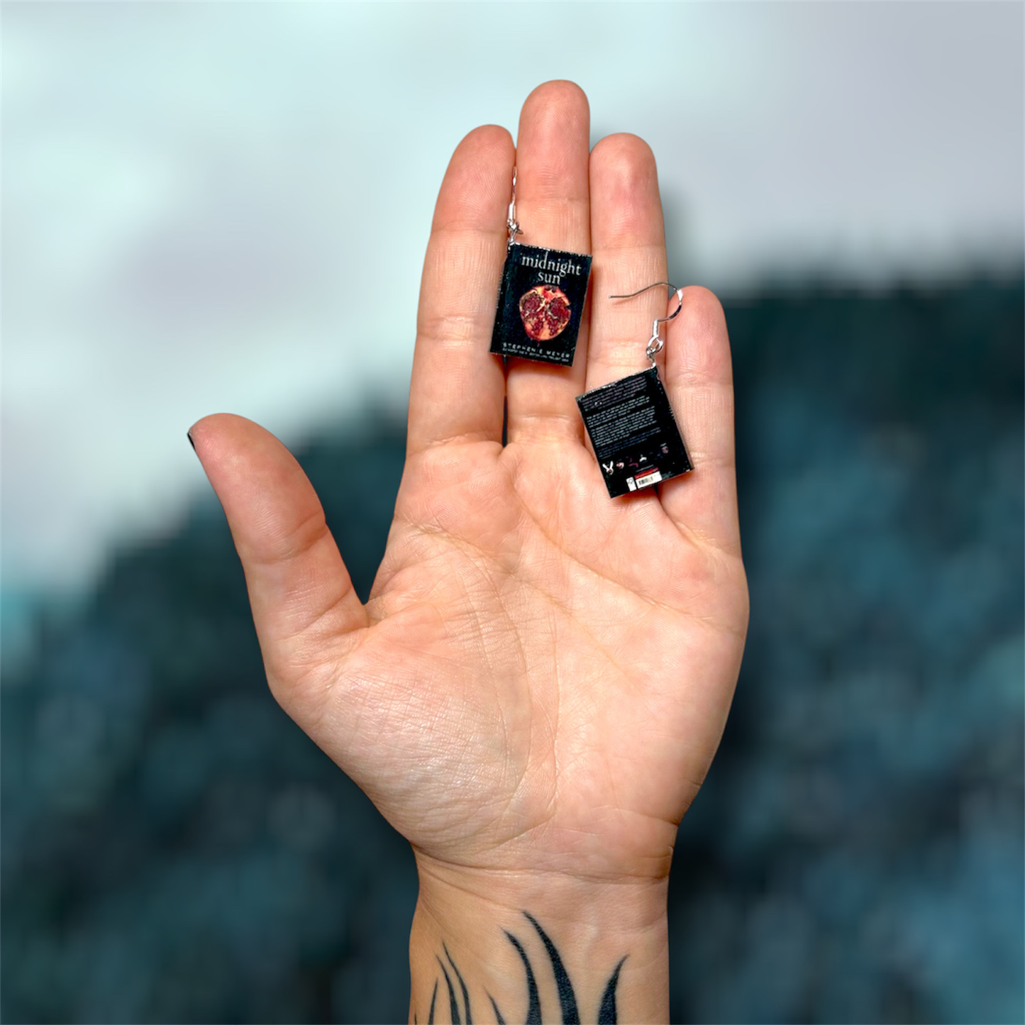 Hand holding a pair of Midnight Sun miniature sparkly vampire book dangle earrings on a blurry blue background