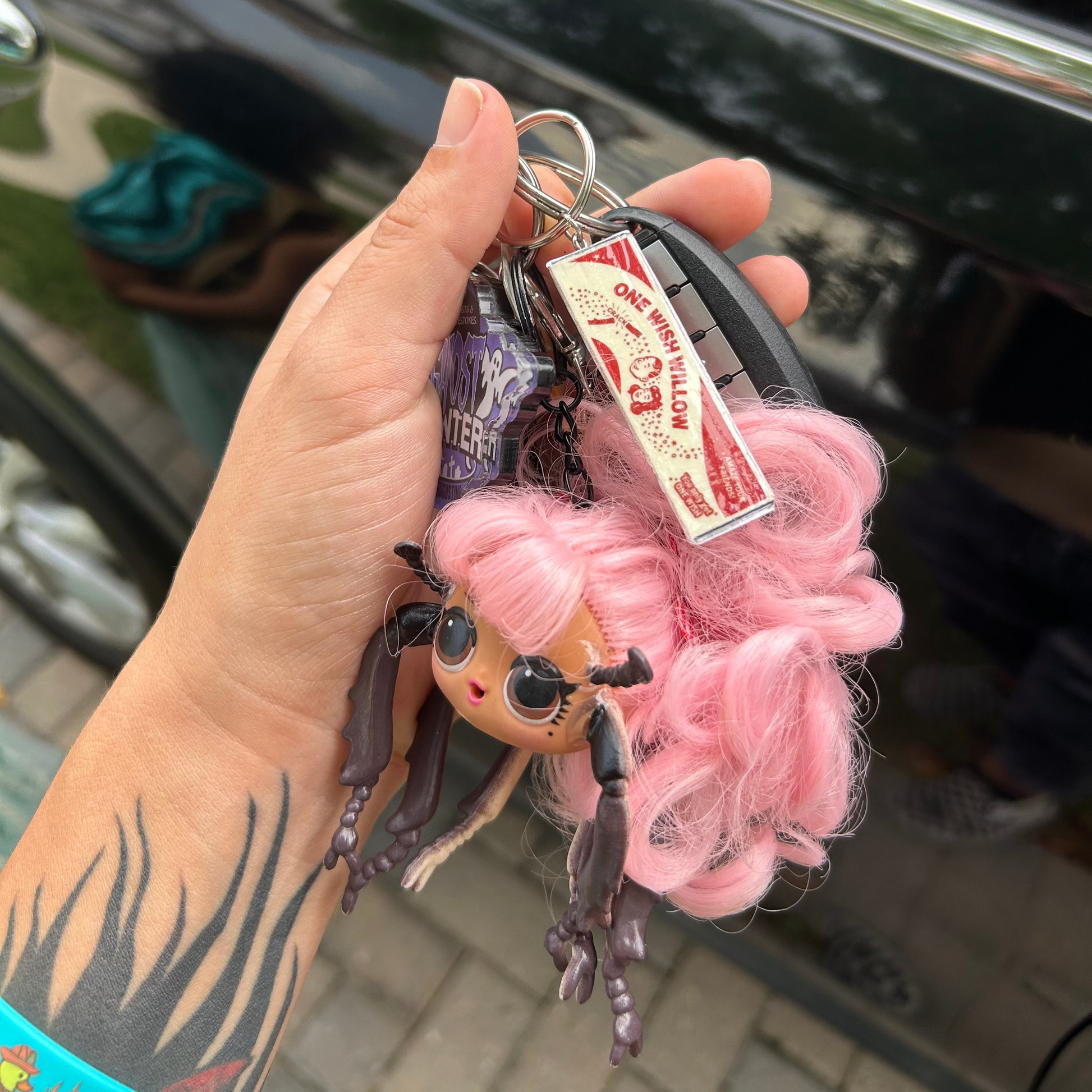 A hand holding a keychain featuring an LOL Surprise doll with insect legs, a Ghost Hunter pendant, and an Obsession-inspired One Wish Willow box replica keychain in front of a blurry car door.