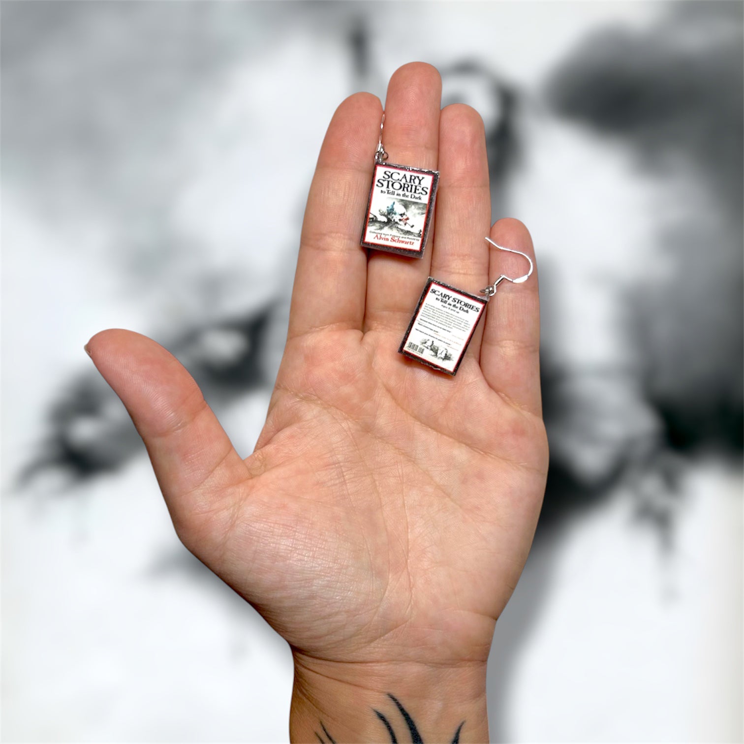 Hand holding a pair of Scary Stories to Tell in the Dark nostalgic horror poetry miniature book earrings on a blurry black and white background