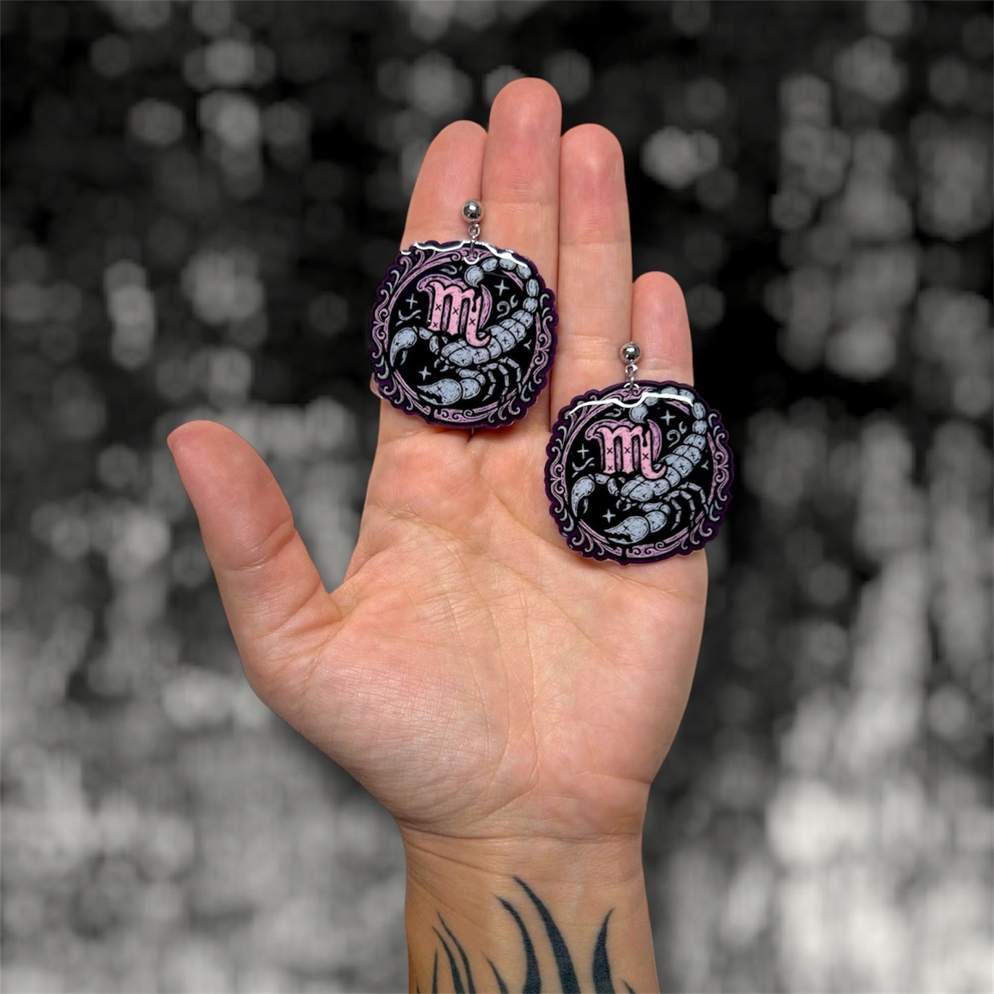 Hand holding a pair of Scorpio zodiac sign scorpion resin dangle earrings on a blurry black and white background