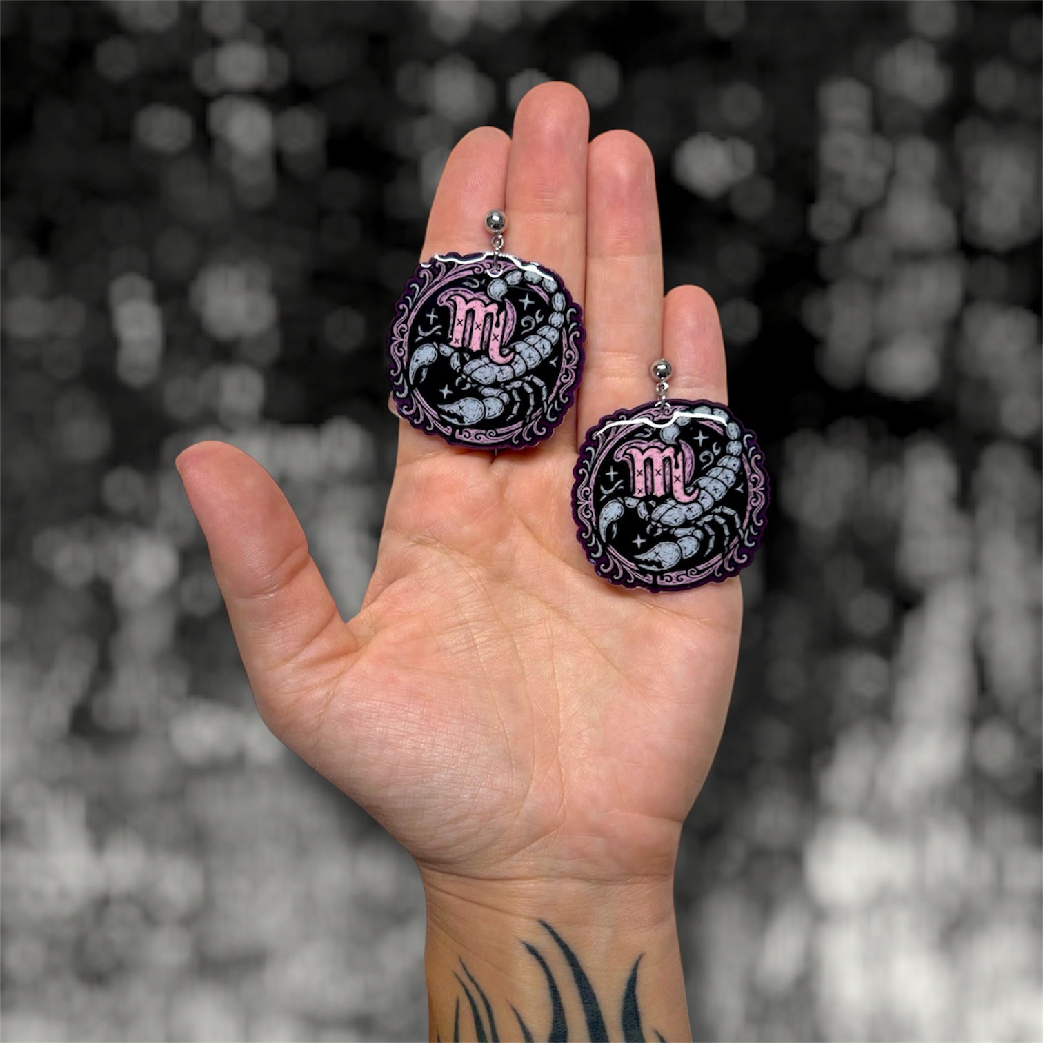 Hand holding a pair of Scorpio zodiac sign scorpion resin dangle earrings on a blurry black and white background