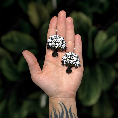 Hand holding human skull flower bouquet spooky resin dangle earrings on a leafy green blurred background