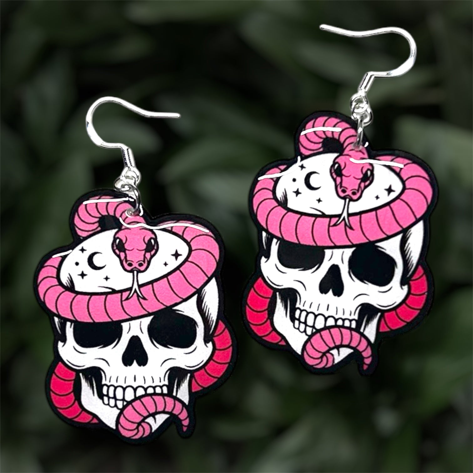 Resin dangle earrings of a pink snake wrapped around a skull with stars and moons, on a blurry green background