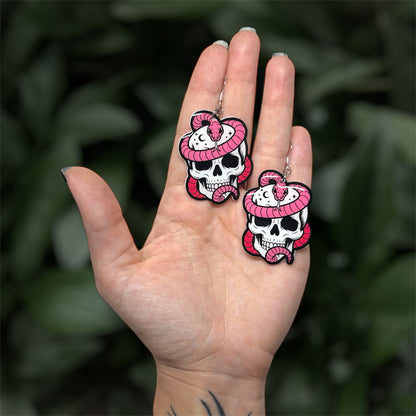 hand holding resin dangle earrings of a pink snake wrapped around a skull with stars and moons, on a blurry green background