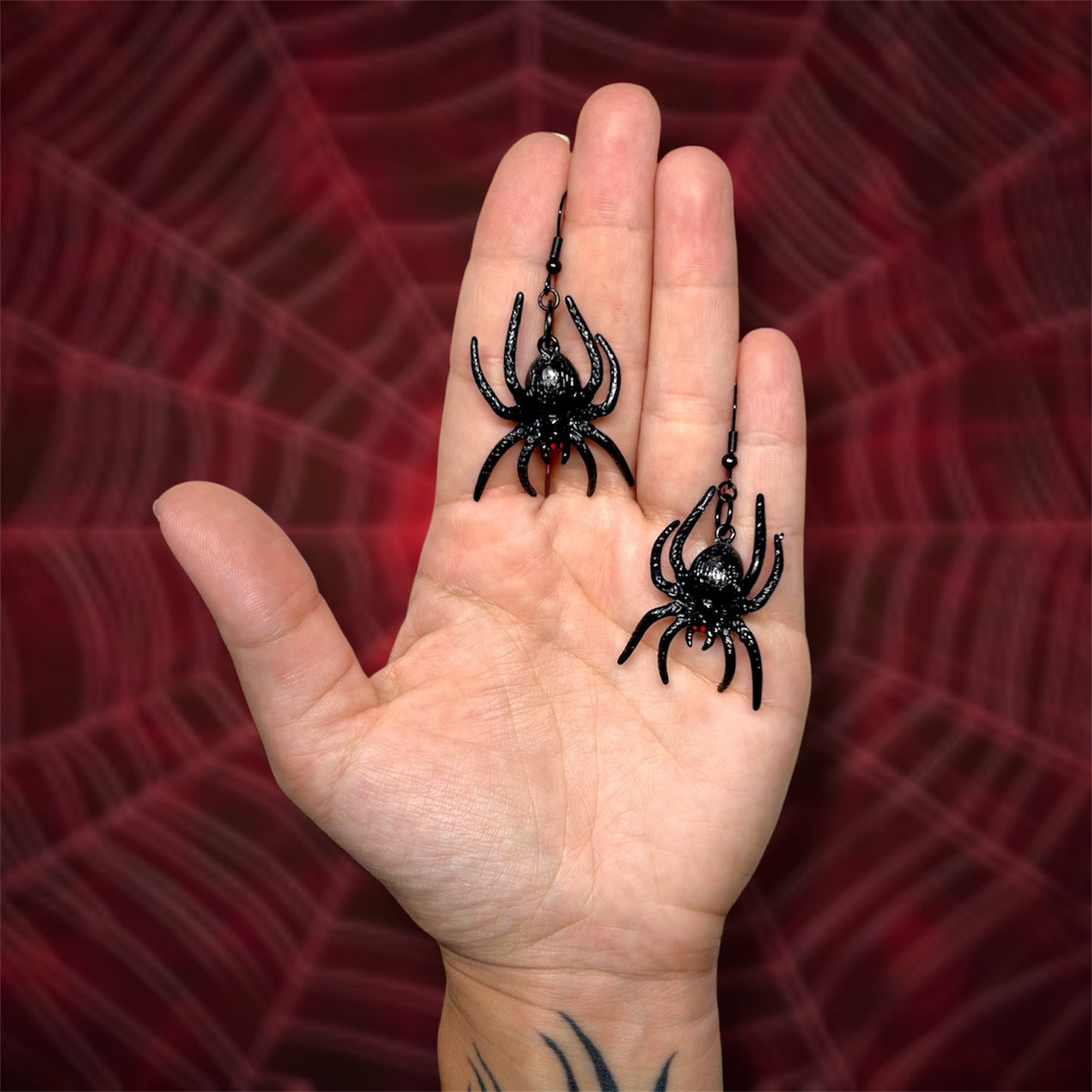 Hand holding a pair of dangle earrings of black spiders with red eyes and fangs on a blurry red spiderweb background