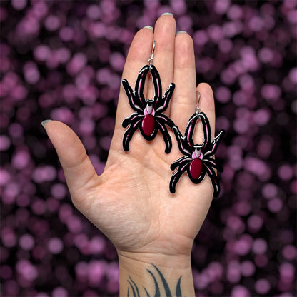 Hand holding a pair of black and pink spider dangle earrings on a blurred purple and black background
