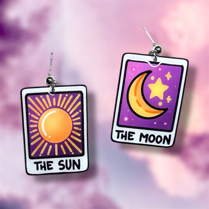 A pair of The Sun and The Moon tarot card witch resin dangle earrings on a blurred purple and pink background