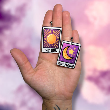 Hand holding a pair of The Sun and The Moon tarot card witch resin dangle earrings on a blurred purple and pink background