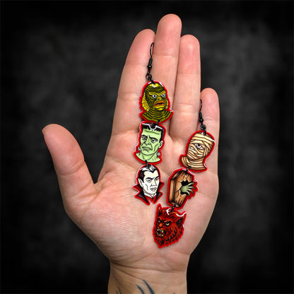 A hand holding a pair of Universal Monsters stacked dangle earrings on a blurred black distressed background